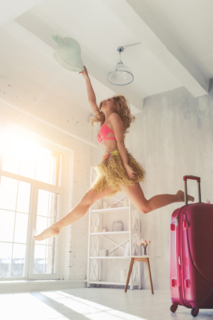 Attractive young woman is preparing for traveling. Dancing in light room in swimsuit, Hawaii accessories and pink sunglasses with big suitcase standing nearby.の写真素材