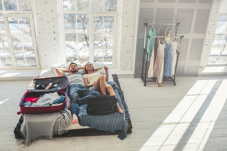 Young romantic couple is preparing for traveling at home. Attractive young woman and handsome man are lying in bed with open travel suitcase. Ready for adventures!の写真素材