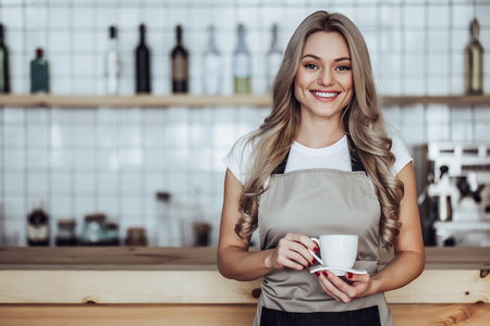 Beautiful female barista is working in coffee shop. Attractive young woman is standing  behind the bar counter, making coffee and welcomes customers.の写真素材