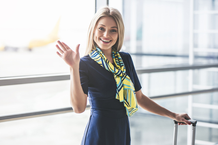 Attractive young female flight attendant is walking in airport terminal and waving handの写真素材
