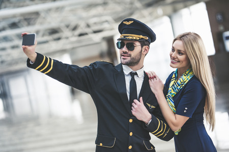 Handsome male pilot and attractive female flight attendant are standing in airport terminal together and making selfie on a smart phone.の写真素材