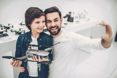 Handsome young man and his cute son are in modern light quadcopter store. Choosing the appropriate drone and making selfie on a smart phone.の写真素材