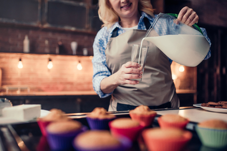 Cropped image of attractive senior woman is cooking on kitchen. Grandmother making tasty baking and pouring milk in the glass.の写真素材