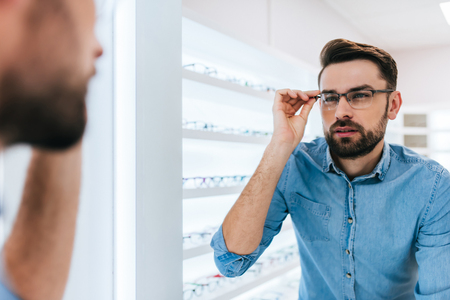 Handsome young man is choosing the most appropriate eyeglasses in modern ophthalmology clinic.の写真素材