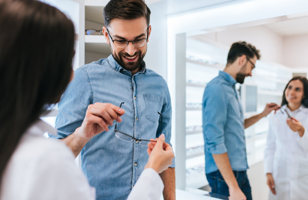 Young female doctor ophthalmologist is helping handsome man to choose the most appropriate eyeglasses. Doctor and patient in modern ophthalmology clinicの写真素材
