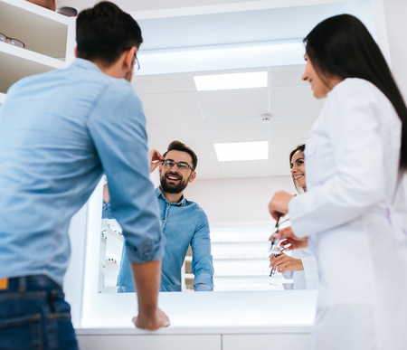Young female doctor ophthalmologist is helping handsome man to choose the most appropriate eyeglasses. Doctor and patient in modern ophthalmology clinicの写真素材