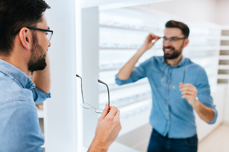 Handsome young man is choosing the most appropriate eyeglasses in modern ophthalmology clinic.の写真素材