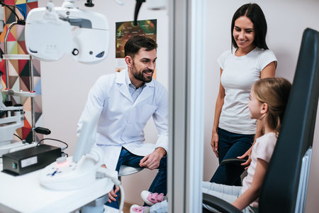 Handsome male doctor  ophthalmologist is checking the eye vision of little cute girl in modern clinic. Mom with daughter in ophthalmology clinic.の写真素材