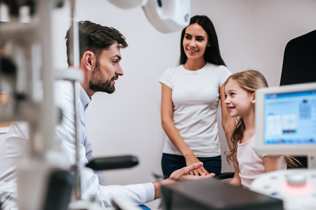 Handsome male doctor  ophthalmologist is checking the eye vision of little cute girl in modern clinic. Mom with daughter in ophthalmology clinic.の写真素材