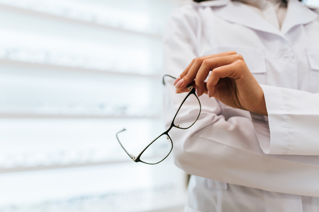 Cropped image of attractive young female doctor in ophthalmology clinic. Doctor ophthalmologist is standing near shelves with different eyeglasses.の写真素材