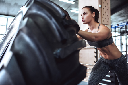 Attractive young sporty woman is working out in gym. Cross fit training. Pushing big tire.の写真素材