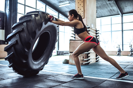 Attractive young sporty woman is working out in gym. Cross fit training. Pushing big tire.の写真素材