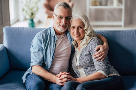Love lives forever! Senior couple at home. Handsome old man and attractive old woman are spending time together. Sitting on a sofa, hugging and holding hands.の写真素材