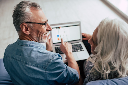 Love lives forever! Senior couple at home. Handsome old man and attractive old woman are spending time together. Sitting on a sofa with laptop.の写真素材