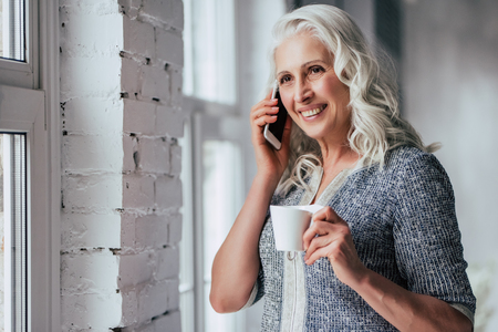 Attractive old woman at home. Senior smiling lady is sitting on a sofa with smart phone in hands while drinking tea.の写真素材