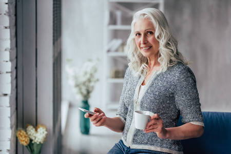 Attractive old woman at home. Senior smiling lady is sitting on a sofa with smart phone in hands while drinking tea.の写真素材