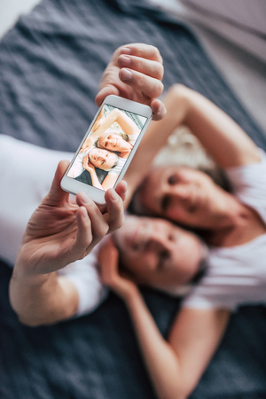 Love lives forever! Senior couple at home. Handsome old man and attractive old woman are enjoying spending time together while lying in bed. Making selfie on a smart phone.の写真素材