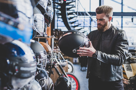 Handsome bearded man in motorcycle shop. Biker is choosing new vehicle and motorcycle accessories. Helmets on wooden background. Safety driving concept.の写真素材