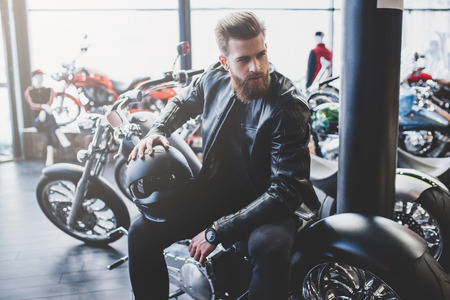 Handsome bearded man in motorcycle shop. Biker is choosing new vehicle and motorcycle accessories. Cropped image of man sittiing with helmet. Safety driving concept.の写真素材