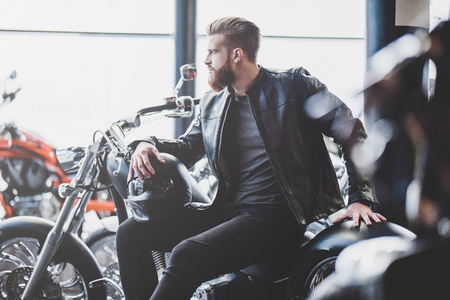 Handsome bearded man in motorcycle shop. Biker is choosing new vehicle and motorcycle accessories. Cropped image of man sittiing with helmet. Safety driving concept.