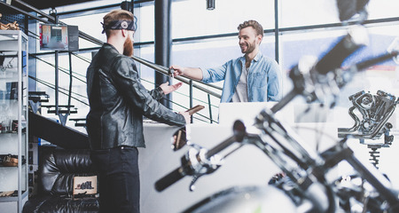 Two men in motorcycle shop. Customer and salesman. Shop assistant is helping bearded man in choosing new vehicle, motorcycle accessories and other biker stuff. Giving keys to new owner.の写真素材