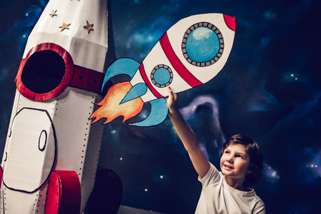 Boy is sitting near toy rocket on space background with drawn small rocket in hand.の写真素材