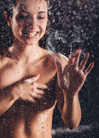 Sexy woman in shower. Attractive young naked woman under water drops isolated on black background is drawing heart on glass.の写真素材