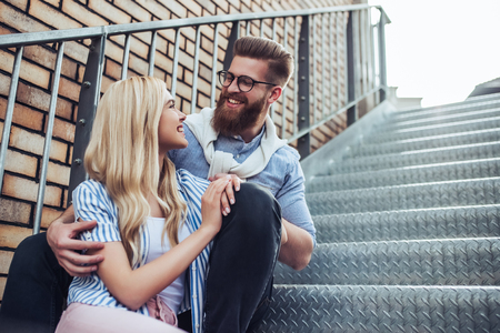 Young stylish happy couple is spending time together outdoors. Attractive woman and handsome bearded man are sitting on stairs in the city.の写真素材