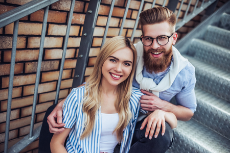 Young stylish happy couple is spending time together outdoors. Attractive woman and handsome bearded man are sitting on stairs in the city.の写真素材