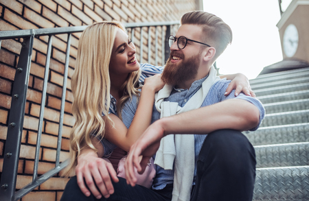 Young stylish happy couple is spending time together outdoors. Attractive woman and handsome bearded man are sitting on stairs in the city.の写真素材