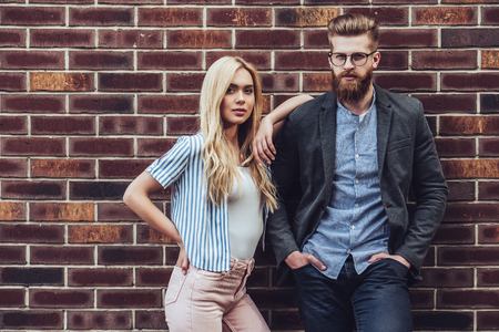 Stylish young couple is spending time together outdoors. Attractive woman and handsome bearded man are posing on brick background.の写真素材