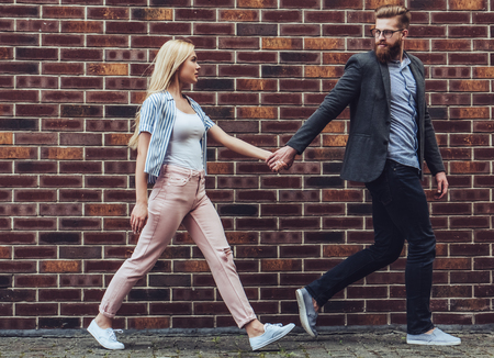 Stylish young couple is spending time together outdoors. Attractive woman and handsome bearded man are walking on brick background and holding hands.の写真素材