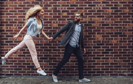 Stylish young couple is spending time together outdoors. Attractive woman and handsome bearded man are walking on brick background and holding hands.の写真素材
