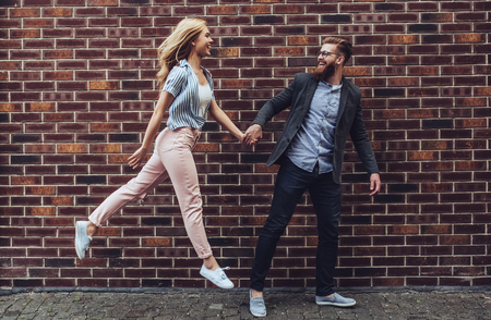 Stylish young couple is spending time together outdoors. Attractive woman and handsome bearded man are walking on brick background and holding hands.の写真素材