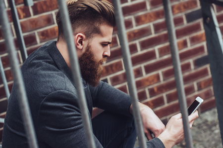 Handsome bearded man is sitting on stairs with smart phone in hands outdoors. Stylish man in the city.の写真素材