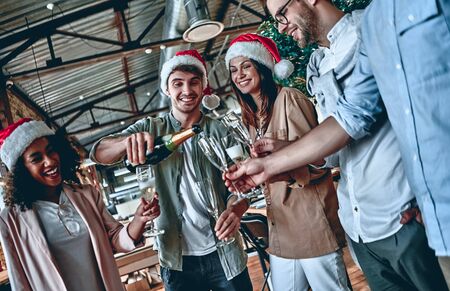 Merry Christmas and Happy New Year 2020!Multiracial young creative people are celebrating holiday in modern office. Group of young business people are drinking champagne in coworking. Successful hipster team of freelancers.の写真素材