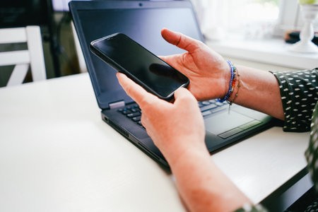 Close-up of a woman's hands holding a smartphone and using a laptop