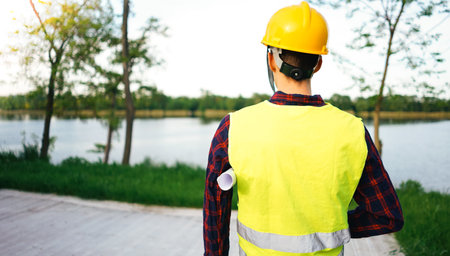 Engineer with yellow helmet and reflective vest standing in front of the lake. Construction worker...