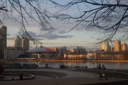 Full color horizontal photo. Evening landscape in the city on the river bank. Sunny weather. Winter has just ended. Trees stand with bare branches. Massive concrete structures and large city buildingsの写真素材