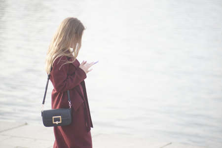 Full color horizontal photo. An unfamiliar beautiful blonde in a beautiful coat is reading something from the phone, standing with her back.の写真素材
