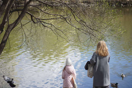 Full color horizontal photo. A parent walks with a girl on a warm sunny spring day in a park on the river bank. People are standing with their backs in outerwear.の写真素材