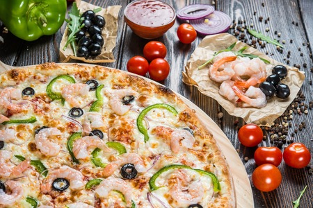 Big pizza with seafood, shrimps and black olives on a round cutting board on a dark wooden background. Food Ingredients. Macroの写真素材