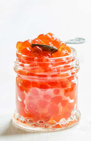Red salmon caviar in a glass jar on a white wooden background, selective focus