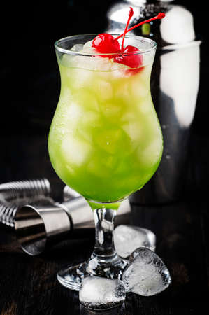 Green alcoholic cocktail with maraschino cherries in hurricane glass, dark bar counter background, selective focusの写真素材