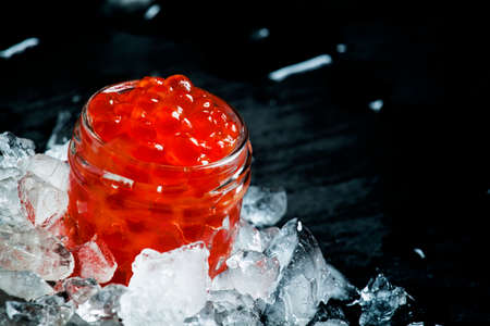 Delicious red caviar in a glass jar on crushed ice on black stone background, selective focusの写真素材