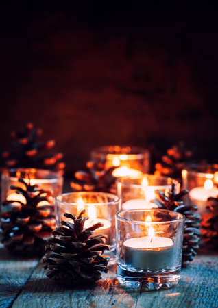 Christmas or New Year composition with burning candles in small glass candlesticks and pine cones, vintage wooden background, festive evening, selective focusの写真素材