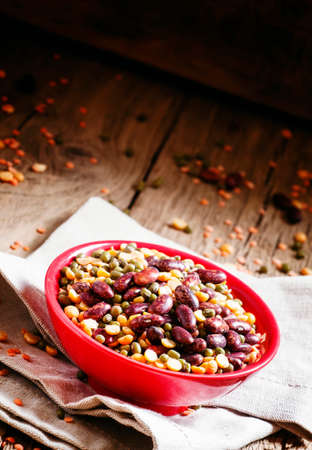 Bean mix: purple beans, green and red lentils, dry peas in a red bowl, vintage wooden background,...