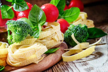 Raw pasta fettuccine, fresh broccoli, green basil leaves, red cherry tomatoes, vintage wooden background, selective focusの写真素材