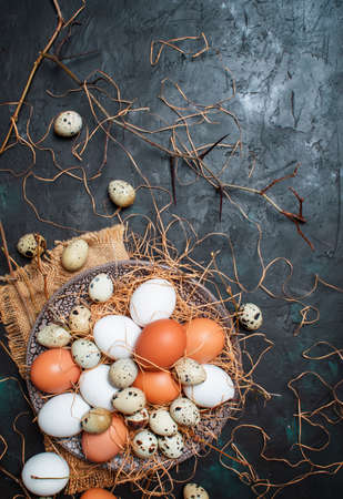Multicolored chicken and quail eggs with straw and branches, spring Easter composition, black...