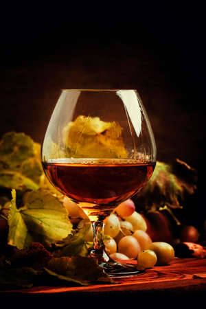 Brandy in a glass and fresh grapes with leaves and a vine, still life in rustic style, vintage wooden background, selective focusの写真素材